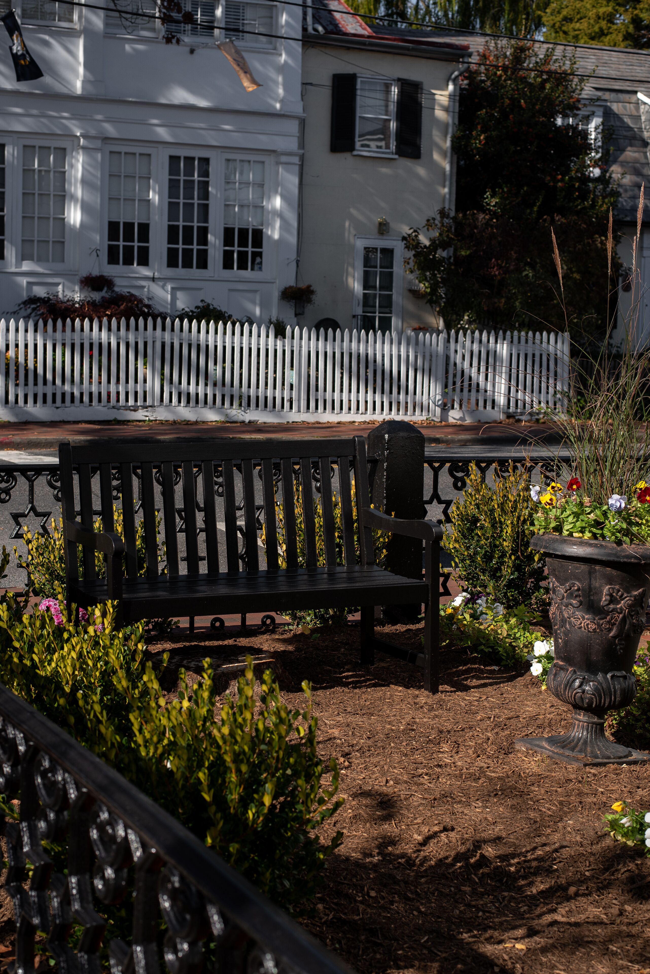 A bench in a garden area with a white picket fence and a building in the background.