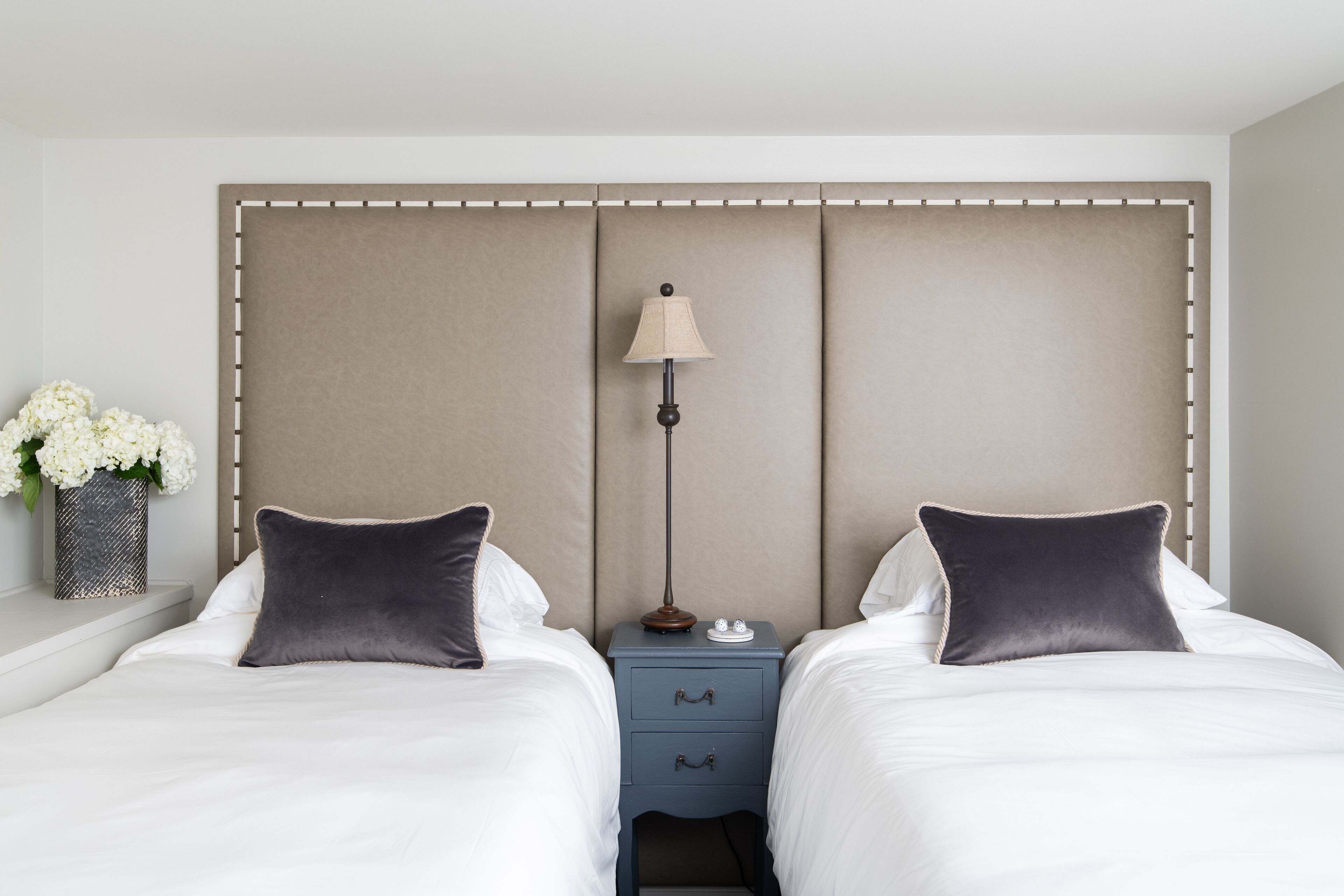A hotel room with two beds, a nightstand with a lamp, and a vase with flowers.