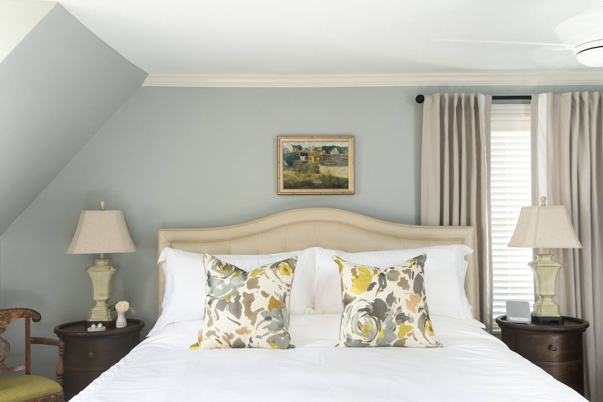 Una cama bien hecha con almohadones florales, un cuadro en la pared y una lámpara a cada lado.
