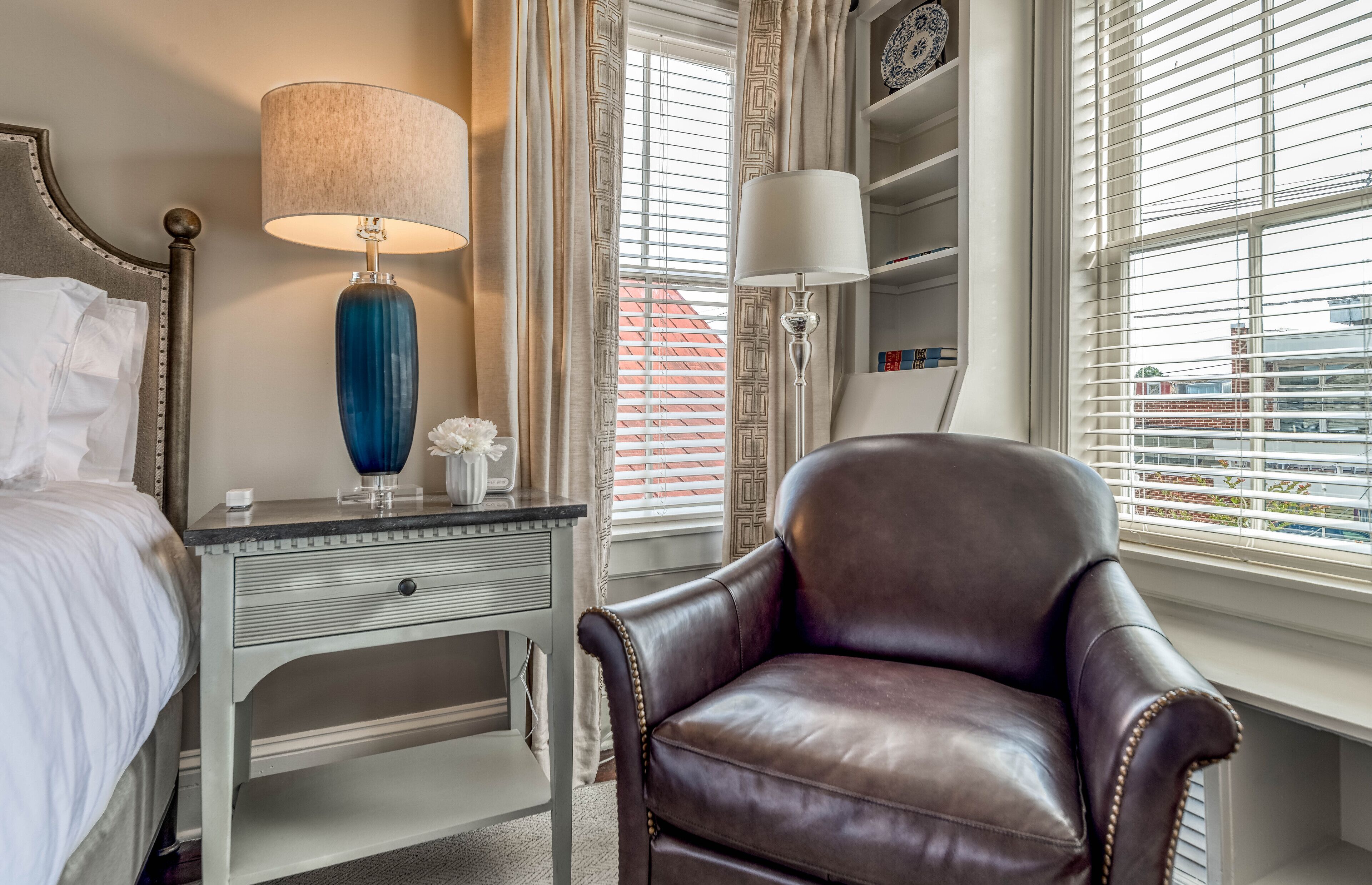 Une chambre à coucher avec un lit, une table de chevet avec une lampe, une chaise et une fenêtre avec des stores.