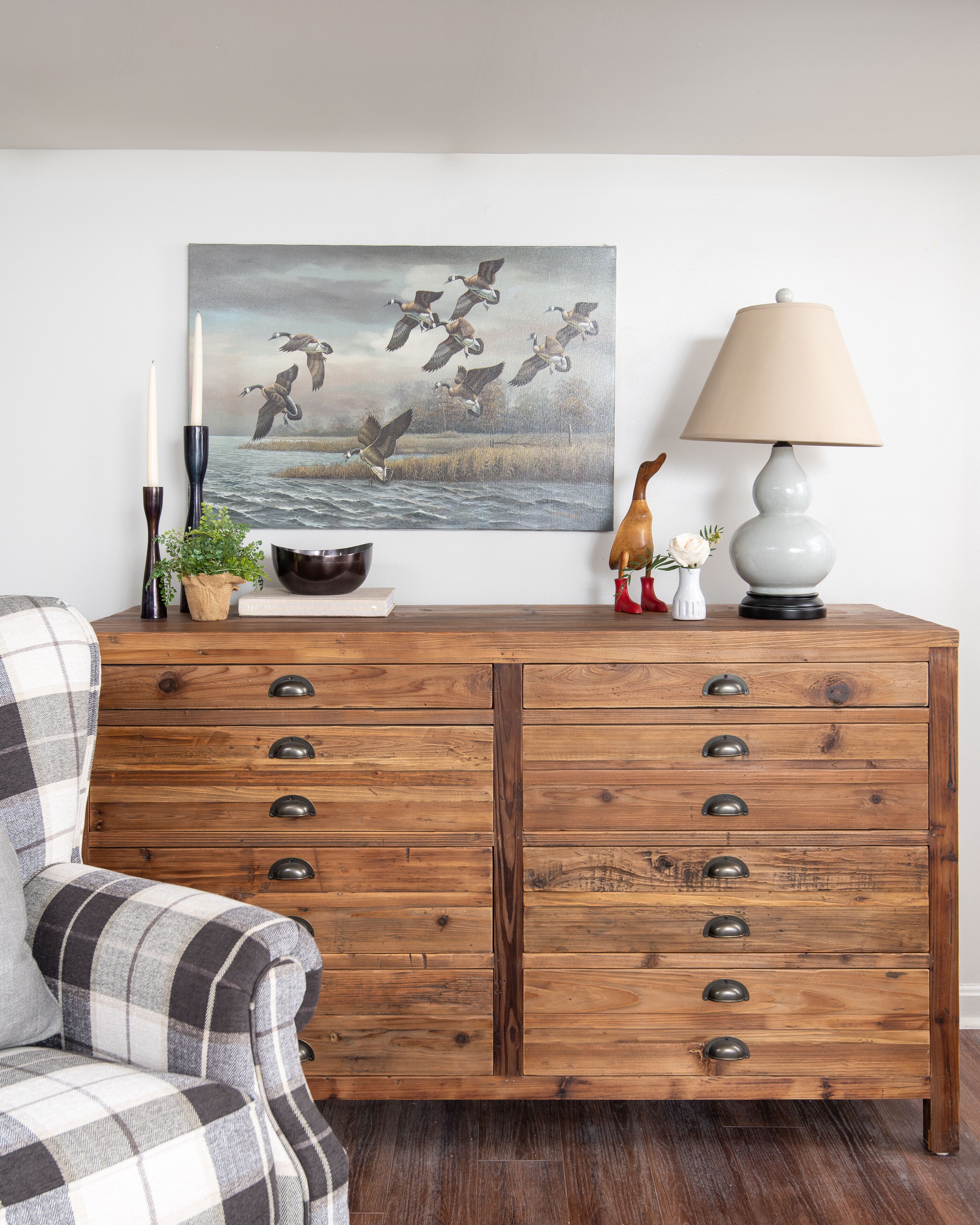A wooden dresser with multiple drawers, a lamp, a painting of birds, a candle, and a small plant.