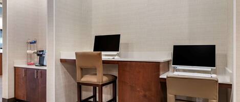 A hotel room with two desks, each with a computer and a chair. There is a small cabinet with a coffee machine and a basket of items.