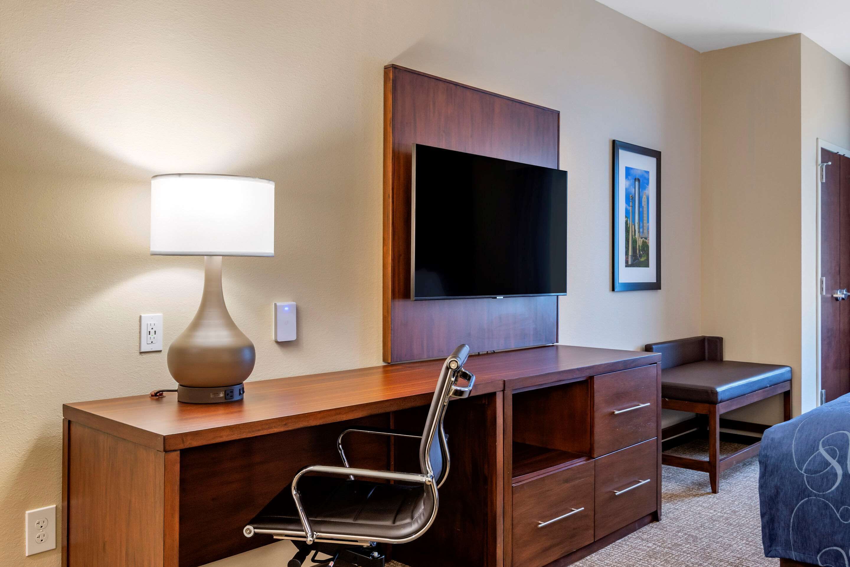 A hotel room with a wooden desk, a chair, a flat-screen TV, a lamp, and a bed.