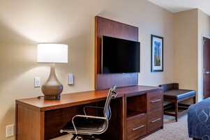 Una habitación de hotel con un escritorio de madera, una silla, un televisor de pantalla plana, una lámpara y una cama.