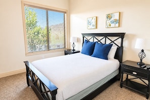 A bedroom with a bed, two bedside tables, a window with blinds, and two wall-mounted paintings. - Comfortable condo with indoor/outdoor pool (Hurricane)