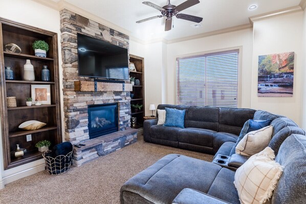 A living room with a stone fireplace, a large flat-screen TV, a dark grey sofa, and a window with blinds. - Comfortable condo with indoor/outdoor pool (Hurricane)