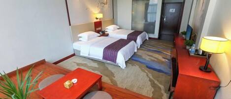 A hotel room with a bed, a desk, a chair, a table, a lamp, and a potted plant.