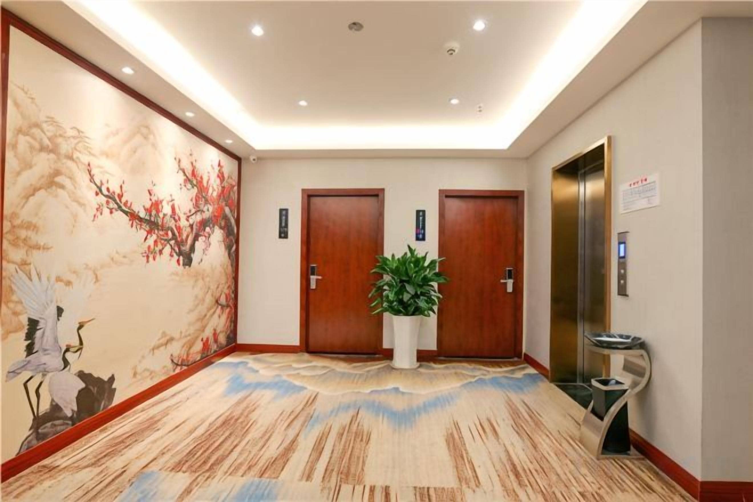 A hotel lobby with a traditional mural, two wooden doors, a potted plant, and an elevator.