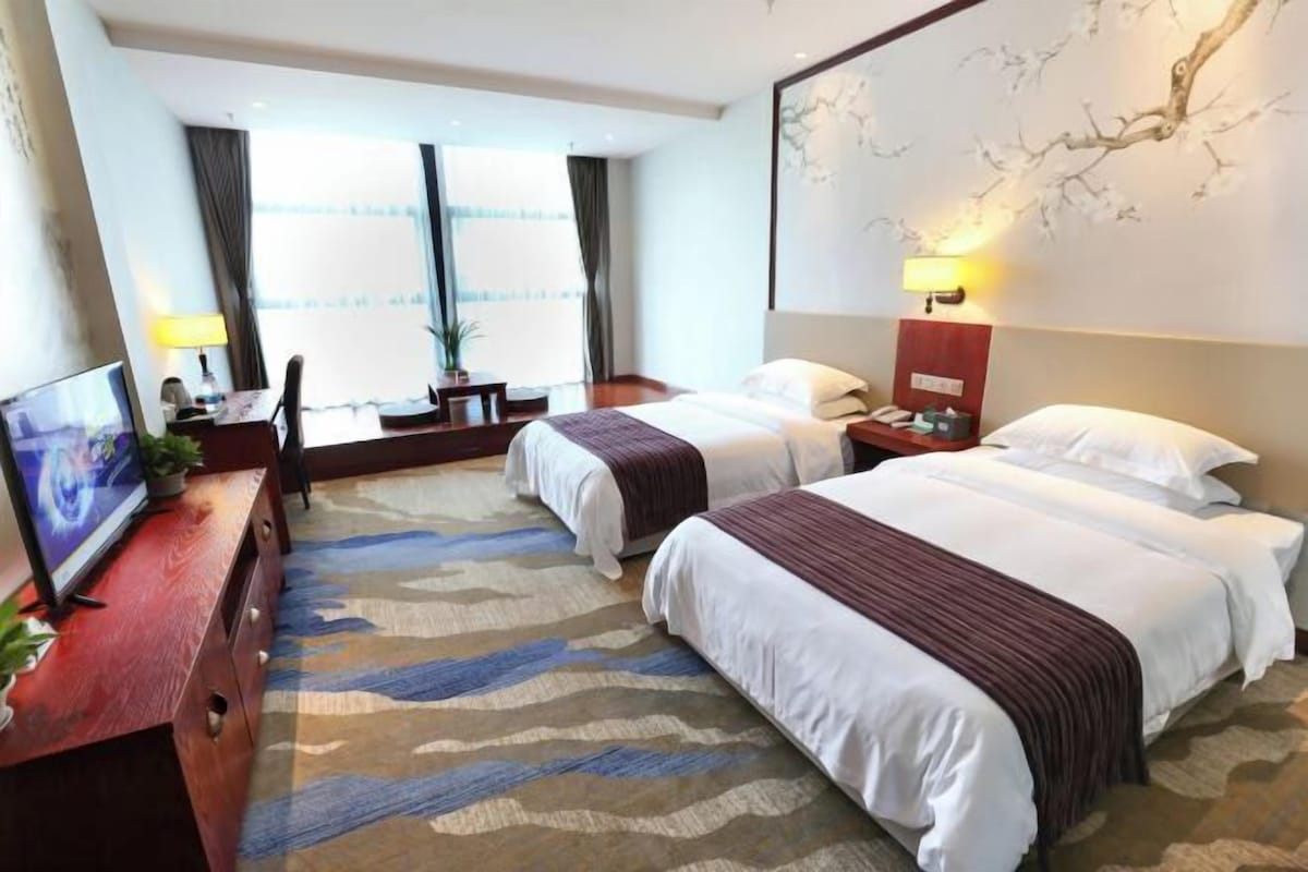 A hotel room with two beds, a desk with a TV, a lamp, and a wall-mounted artwork.