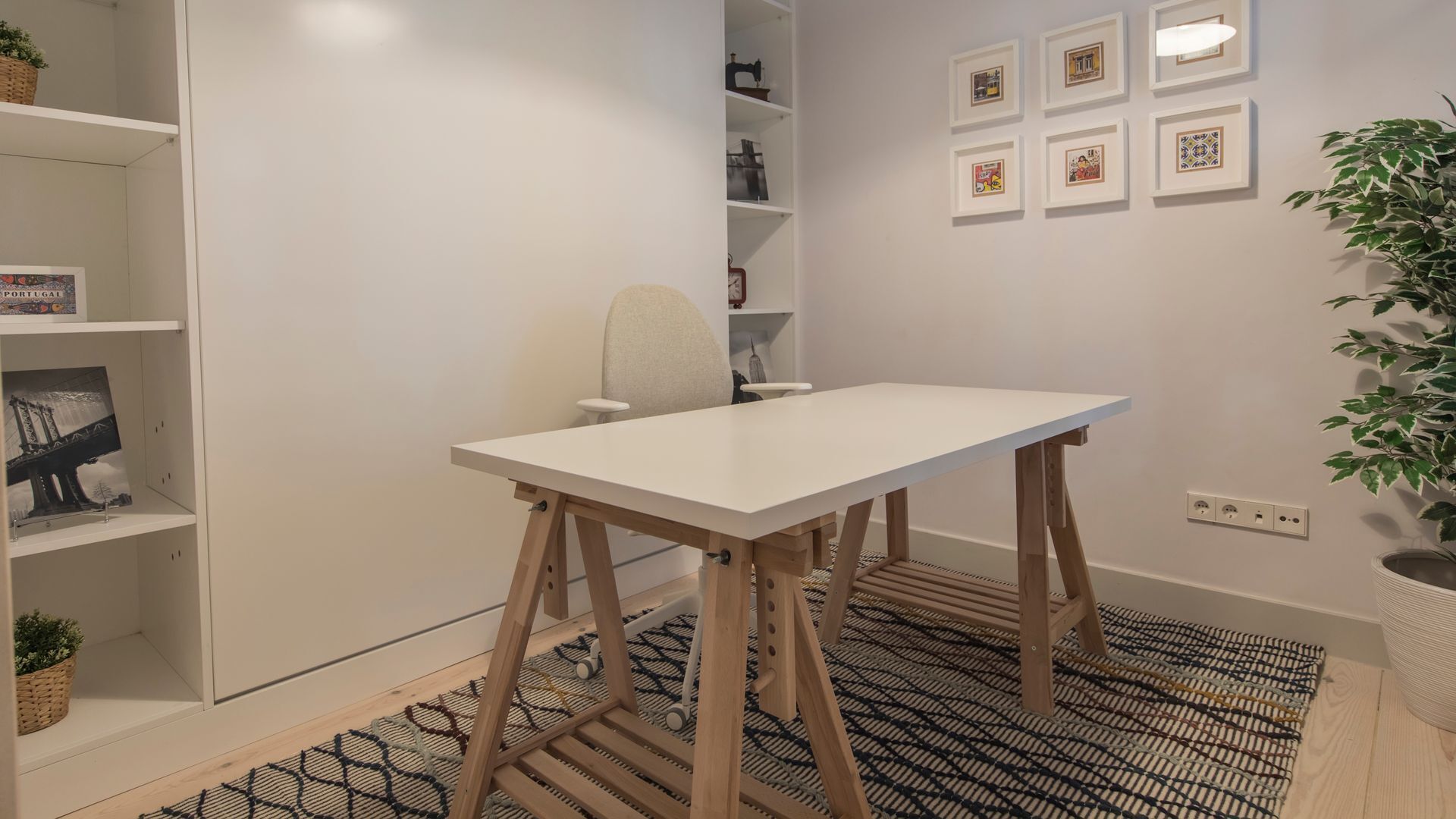 A room with a white table, a chair, a shelf with a book, and a wall with framed pictures.