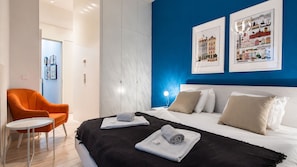 A hotel room with a bed, a chair, a bedside table, and artwork on the wall.