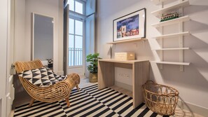 Une pièce avec un sol à rayures noires et blanches, une chaise en osier agrémentée d’un coussin à motifs géométriques, un bureau avec une plante et un tableau encadré accroché au mur.