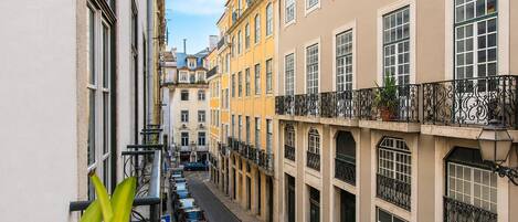 A narrow street flanked by multi-story buildings with balconies, parked cars, and a clear sky.