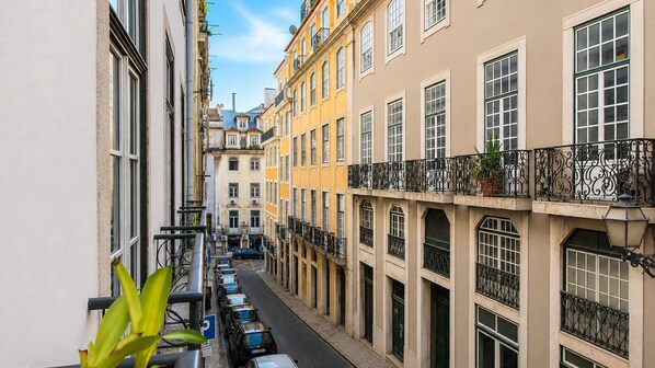 A narrow street flanked by multi-story buildings with balconies, parked cars, and a clear sky.
