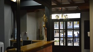 Un mostrador de recepción con encimera de madera, una planta colgante y vistas al exterior a través de una puerta de cristal con la etiqueta 'SALIDA'.