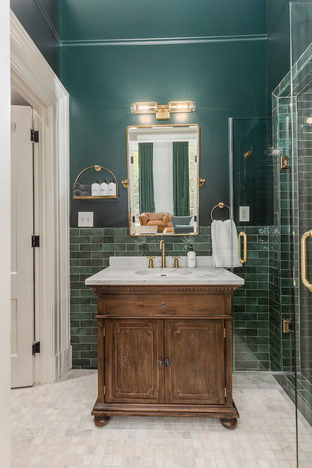 A bathroom with a dark green tiled wall, a wooden vanity with a mirror, and a glass shower enclosure.