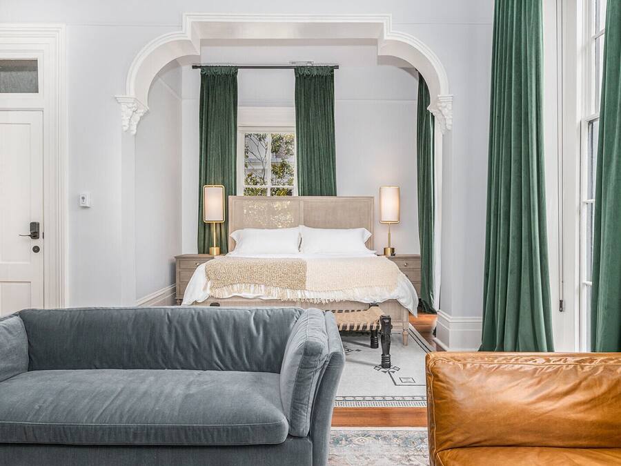 A bedroom with a large bed, two bedside tables, green curtains, a grey sofa, and a brown leather chair.