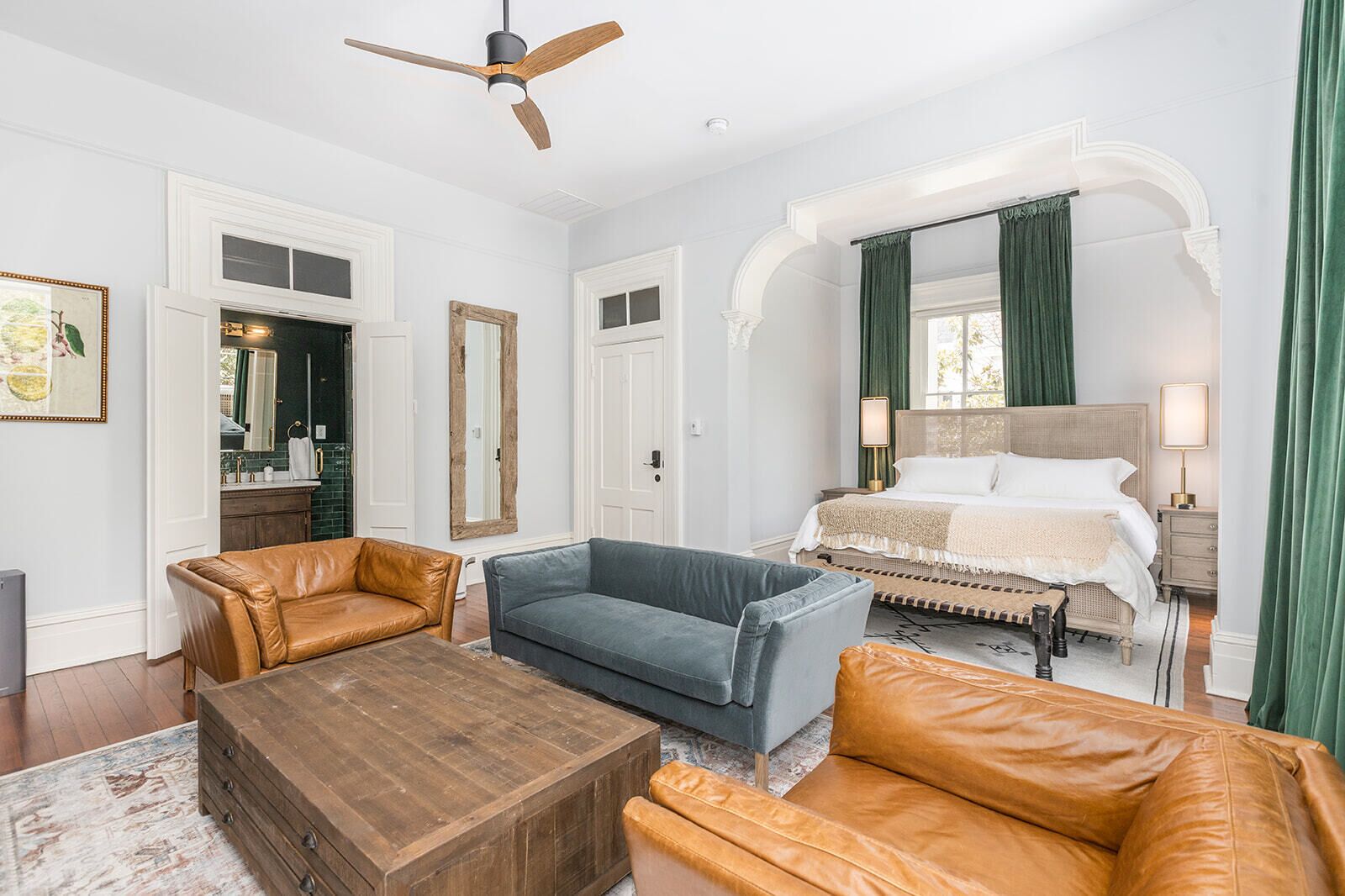 A hotel room with a bed, two sofas, a wooden coffee table, a ceiling fan, and a large mirror.