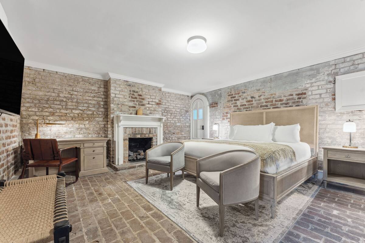 A bedroom with a brick wall, a bed, a desk, a chair, a fireplace, and a nightstand.