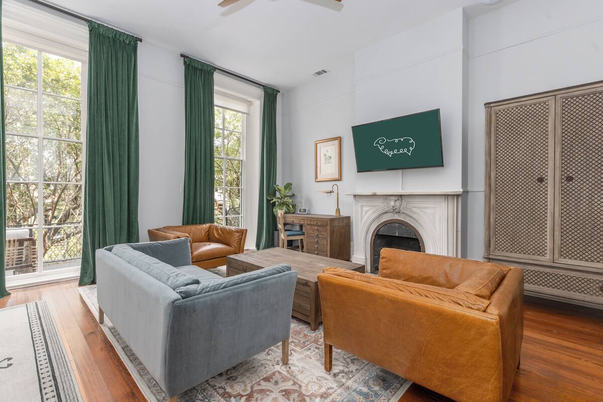 A living room with a sofa, armchair, and coffee table. There is a fireplace, a TV mounted on the wall, and a large window with curtains.