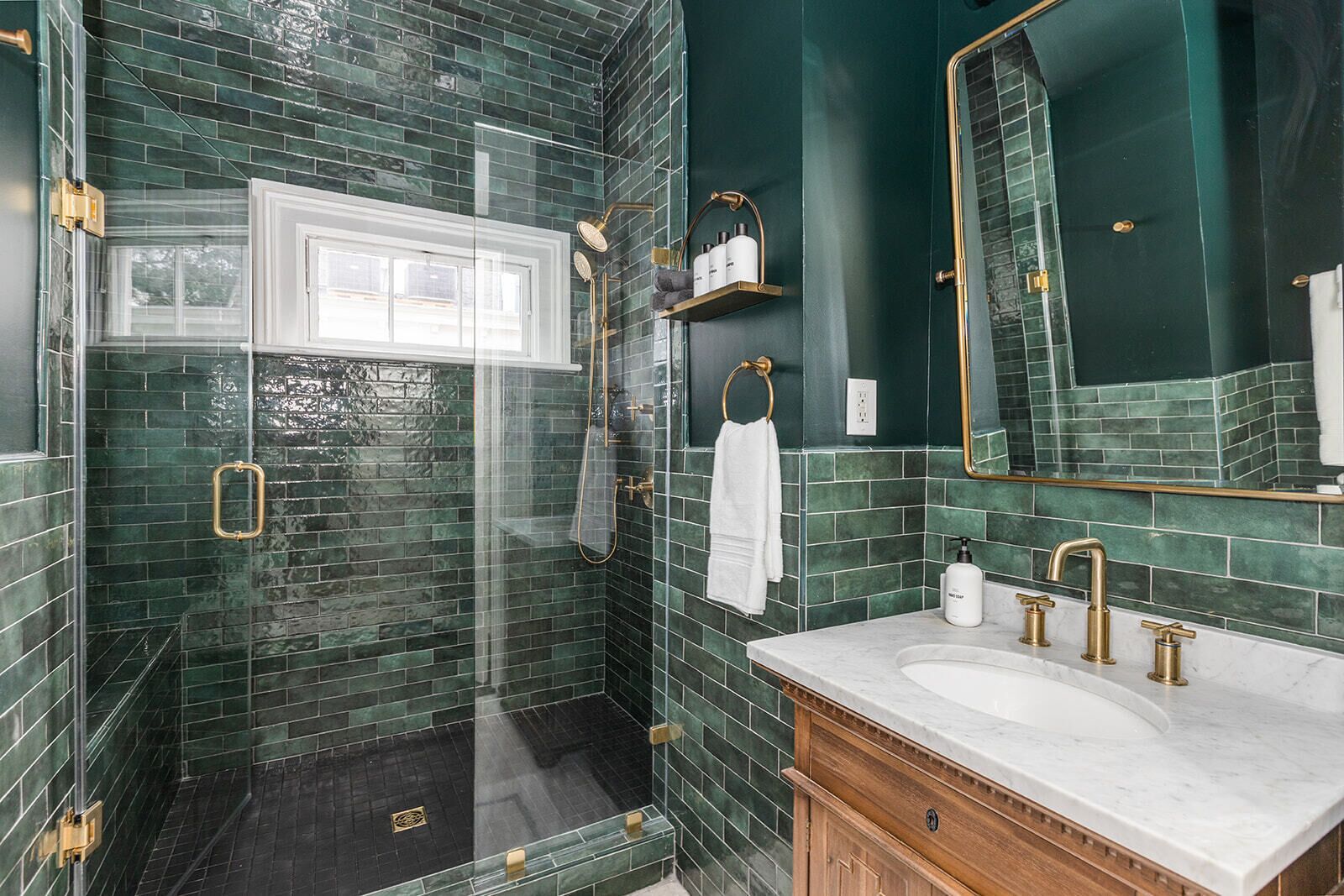 A bathroom with dark green tiles, a glass shower enclosure, a white vanity with gold fixtures, and a large mirror.
