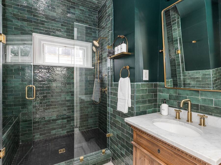 A bathroom with dark green tiles, a glass shower enclosure, a white vanity with gold fixtures, and a large mirror.