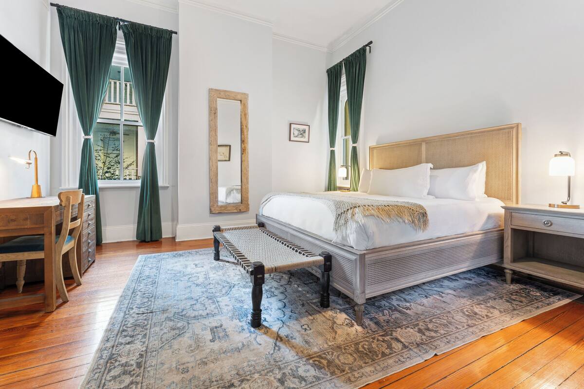 A hotel room with a bed, bedside table, lamp, chair, desk, TV, and a large window with curtains.