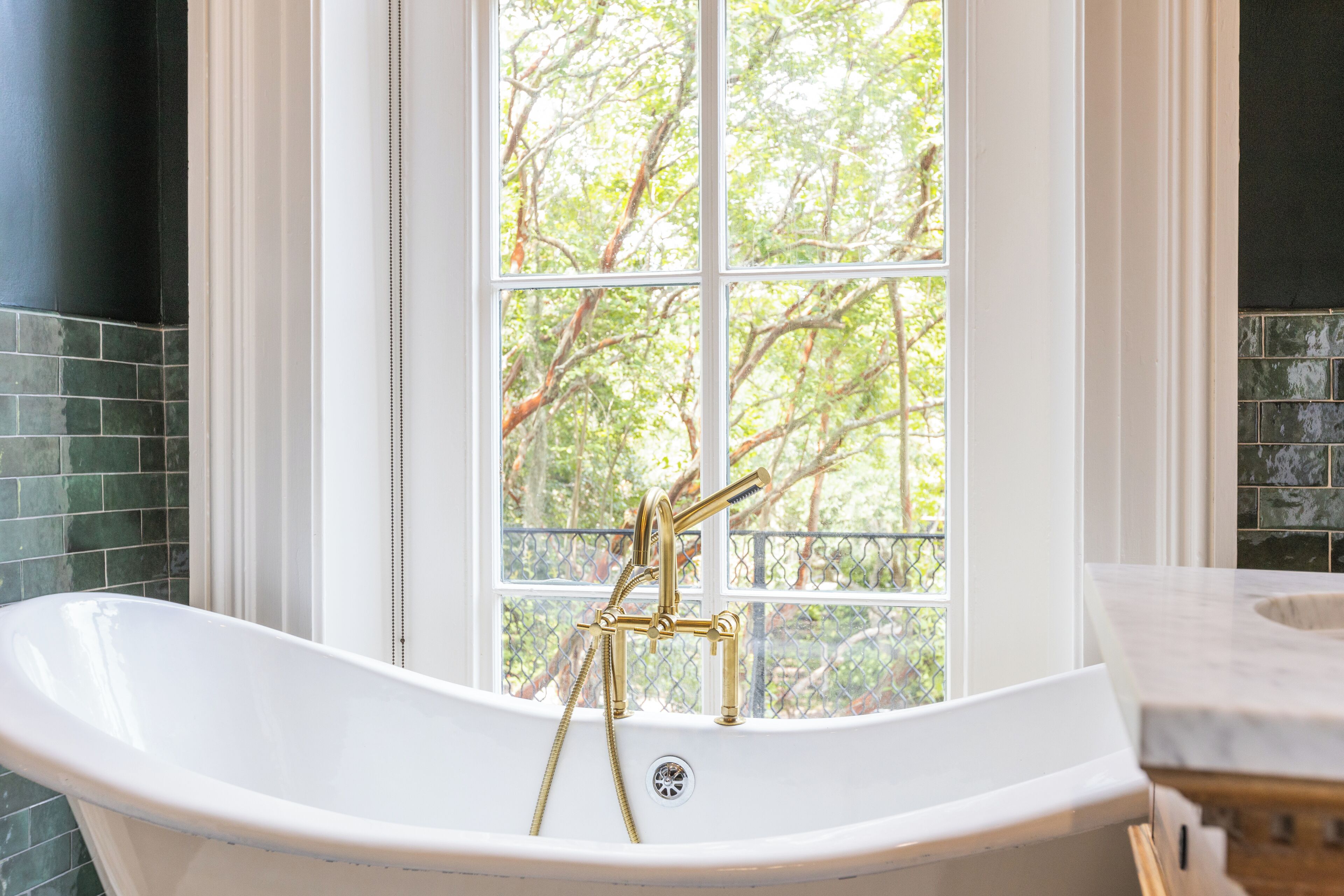 A freestanding bathtub with a gold faucet and shower head, positioned in front of a window with a view of trees.