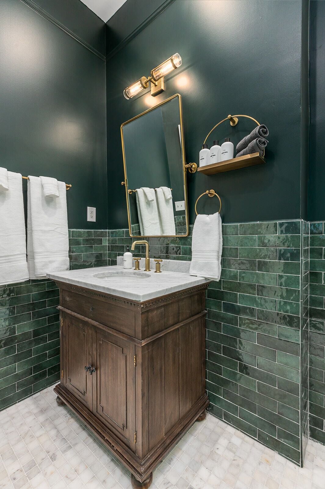A bathroom with a dark green tiled wall, a wooden vanity with a white sink, and a mirror with a gold frame.