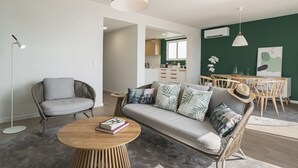 Una sala de estar moderna con un sofá gris, una mesa de centro redonda de madera y un sillón de mimbre. Se observa un comedor con sillas de madera y una mesa al fondo.