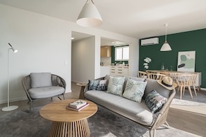 A modern living room with a grey sofa, a round wooden coffee table, and a wicker armchair. A dining area with wooden chairs and a table is visible in the background.