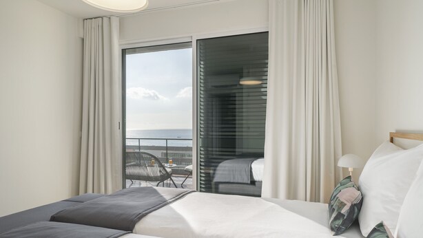 Habitación de hotel con una cama, una silla, una mesa y vistas al mar a través de un amplio ventanal.