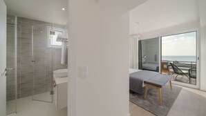 Un dormitorio moderno con una cama grande, un pequeño banco, cabina de ducha de vidrio y un balcón con vistas al mar.