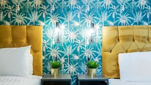 A hotel room with two beds, each with a gold headboard and white bedding. There are two small black nightstands with gold lamps and potted plants. The wall features a tropical leaf pattern. - Queen Street Apartments by Sasco (Blackpool)