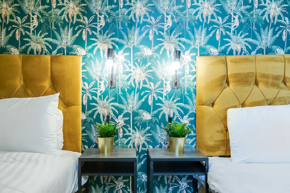 Een hotelkamer met twee bedden, elk met een gouden hoofdeinde en witte beddengoed. Er staan twee kleine, zwarte nachtkastjes met goudkleurige lampen en potplanten. De wand heeft een tropisch bladmotief.