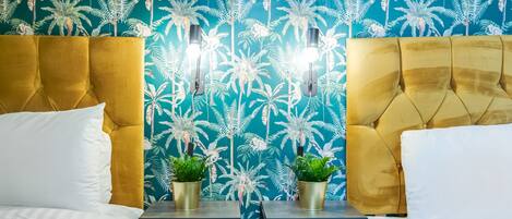 Habitación de hotel con dos camas, cada una con cabecero dorado y ropa de cama blanca. Hay dos mesitas de noche negras con lámparas doradas y plantas en macetas. La pared presenta un diseño con hojas tropicales.