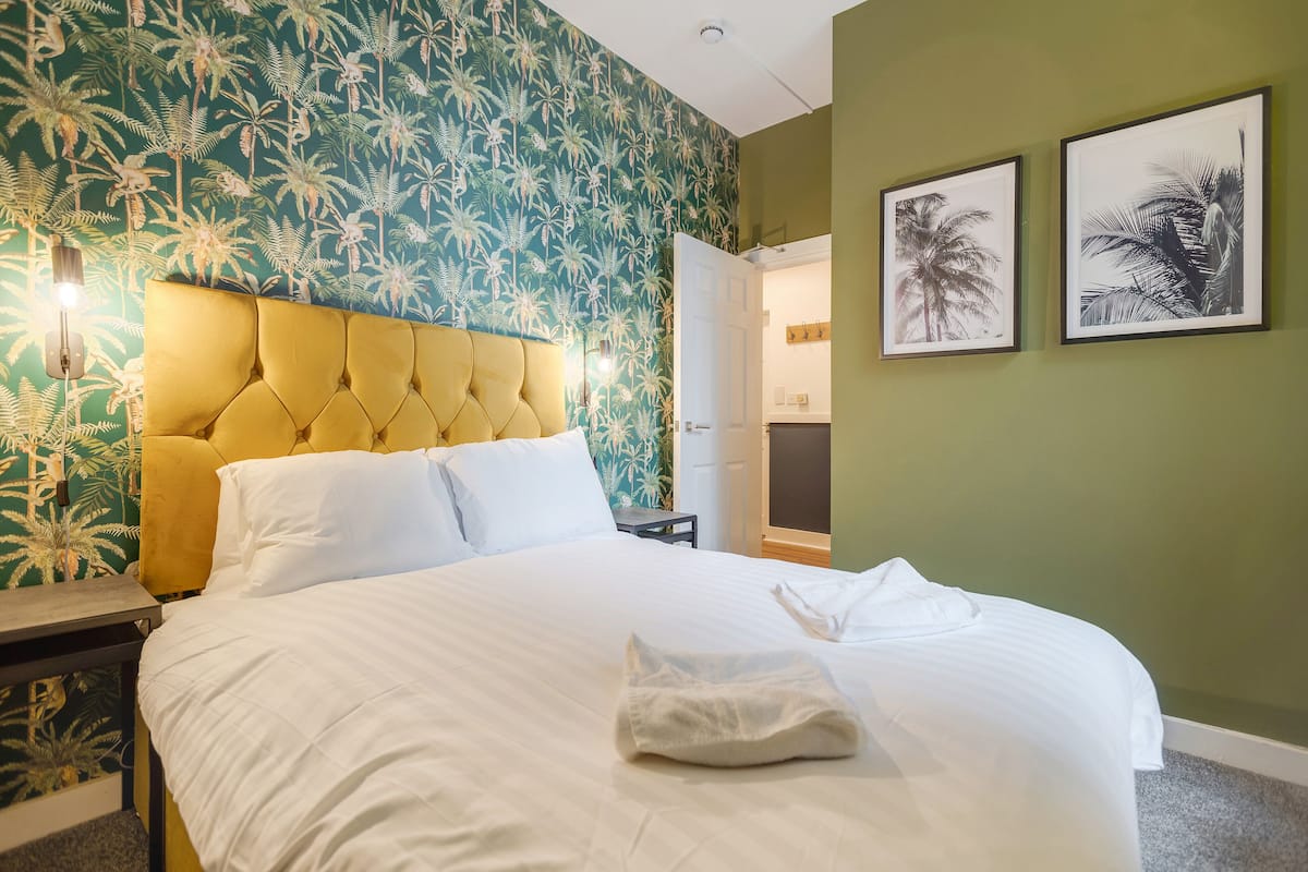Een hotelkamer met een bed, hoofdeinde, nachtkastje, een muur met tropische behang en twee ingelijste foto’s aan de wand.
