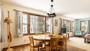 Un comedor con mesa y sillas de madera, una araña y grandes ventanales con vistas a árboles.