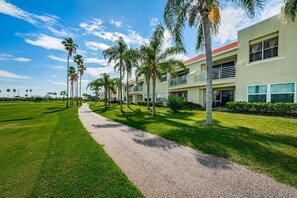 A pathway leading to a building with multiple windows and a red roof, surrounded by palm trees and a well-maintained lawn.