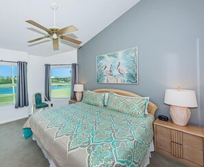 A bedroom with a bed, a ceiling fan, a nightstand, a lamp, a chair, and a window with curtains.