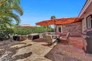 An outdoor patio with stone flooring, a dining table with chairs, a grill, and orange umbrellas.