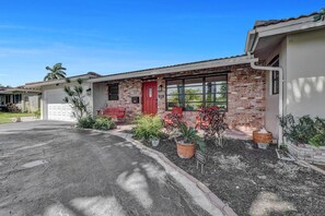 A single-story house with a red door, brick facade, and a front yard with potted plants.