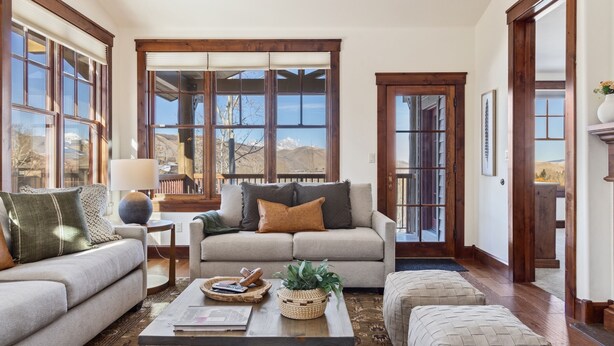 A cozy living room with a beige sofa, a coffee table with books and a plant, and large windows with a mountain view. - Outpost: Grand View 3230 - Air Conditioning! (Jackson)