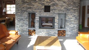 Ein Wohnzimmer mit Kamin aus Stein, Holzmöbeln und Blick ins Freie.