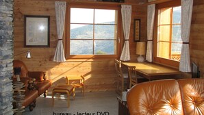 Ein gemütliches Zimmer mit Holzwänden, einem Schreibtisch, einem Stuhl, einer Couch und einem Fenster mit Blick auf die Berge.