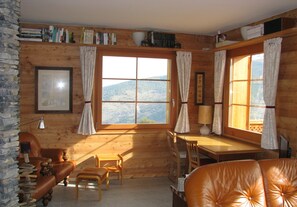 Ein gemütliches Zimmer mit Holzwänden, einem Schreibtisch, einem Stuhl, einer Couch und einem Fenster mit Blick auf die Berge.