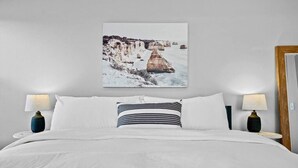 Een slaapkamer met een groot bed, twee nachtkastjes met lampen, en een lijst met een afbeelding van een kustlandschap aan de muur.