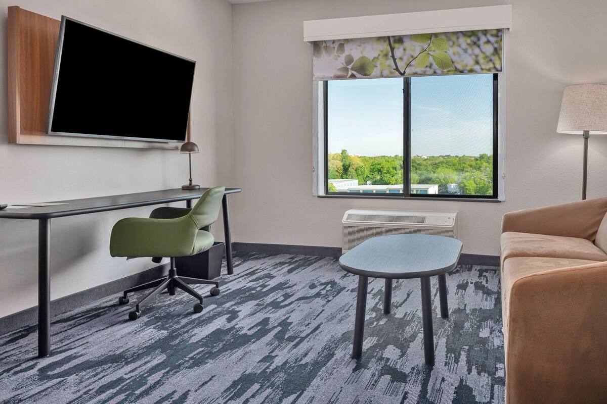 Une chambre d’hôtel moderne équipée d’un téléviseur à écran plat fixé au mur, d’un bureau avec une chaise, d’une petite table ronde, d’un canapé et d’une grande fenêtre donnant sur un paysage verdoyant et des bâtiments.