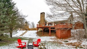 Una cabaña con chimenea de piedra, terraza de madera y suelo cubierto de nieve.