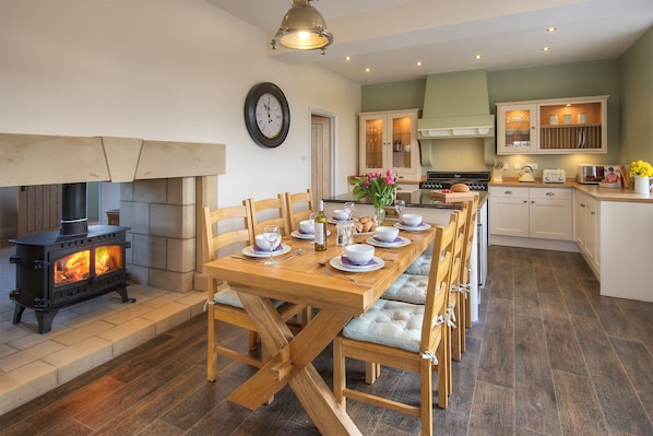A cozy dining area with a wooden table set for six, a wood-burning stove, and a kitchen in the background.